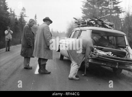 Fotografia in bianco e nero di quattro uomini, ciascuno indossando un cappotto o una giacca, in piedi su un vuoto del tratto di strada con una stazione di sosta carro che ha i corpi di diversi cervi nel suo overhead rack; un uomo in primo piano a destra sembra cambiare un pneumatico posteriore mentre due uomini guardano il quarto uomo film o fotografie il procedimento da sfondo a sinistra; da conifere e altri alberi visibile sullo sfondo, fotografato durante una battuta di caccia e viaggio di pesca si trova in Alaska, Stati Uniti d'America, 1955. () Foto Stock