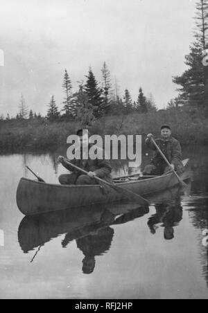 Fotografia in bianco e nero di due persone di mezza età uomini, ciascuno indossa una giacca imbottita e un cappuccio, seduti in canoa con pale nelle loro mani con i loro riflessione visibile in acqua sotto la barca e alberi di conifere sulla riva sullo sfondo, fotografato durante una battuta di caccia e viaggio di pesca si trova in Alaska, Stati Uniti d'America, 1955. () Foto Stock