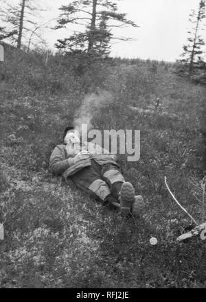 Fotografia in bianco e nero di un uomo di mezza età, in piena vista di lunghezza, che indossa una giacca, tute, stivali e un cappuccio, sdraiato, con gli occhi chiusi, su un dolcemente classificato, Grassy Slope; una mano è nella sua tasca della giacca, l'altra tiene una sigaretta, e recentemente il fumo espirato sorge sopra la sua testa; con numerosi alberi di conifere visibile sullo sfondo, fotografato durante una battuta di caccia e viaggio di pesca si trova in Alaska, 1955. () Foto Stock