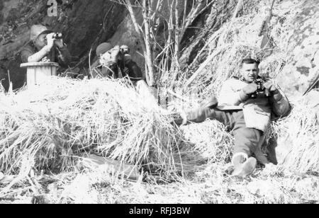 Fotografia in bianco e nero di tre persone di mezza età gli uomini in un ambiente esterno con erbe secche e rami sul terreno e una collina rocciosa o cliff in background; due uomini a sinistra usare il binocolo, un terzo uomo a destra, indossando una calda giacca di caccia, tall lace-up degli stivali e guanti, reclinabile sul terreno tenendo il suo binocolo lontano dal suo volto, fotografata durante una battuta di caccia e viaggio di pesca si trova in Alaska, 1955. () Foto Stock