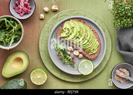Sandwich di Avocado e insalata verde con cubetti di prosciutto su marrone-verde sfondo testurizzato, vista dall'alto con ingredienti intorno Foto Stock