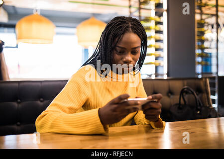 Giovane donna africana presso il cafe a bere caffè e utilizzando il telefono cellulare Foto Stock