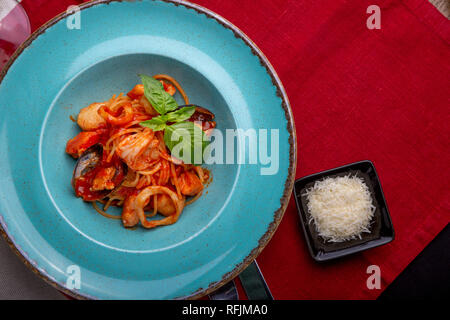 Cucina Italiana. Pasta con pesce e frutti di mare, i pomodori e il formaggio in un blu, una forchetta e un coltello su uno sfondo luminoso. Foto Stock