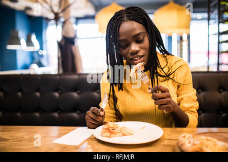 Giovani afro american donna che indossa in maglione giallo mangiare la pizza in cafe Foto Stock