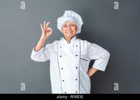 Senior chef donna studio isolato permanente su grigio in posa mostra ok allegro Foto Stock