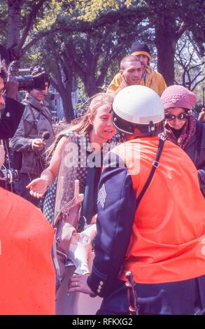 Fotografia a colori che mostra una scena dal 1971 giorno di maggio proteste situato in Washington, DC; un giovane biondo, femmina antiwar protestor, indossando una marina e bianco abito senza maniche, curve a litigare con un motociclo poliziotto che indossa un casco bianco e un giubbotto di colore arancione, con numerosi giornalisti (uno tenendo un microfono, altri con le telecamere) un'altra donna, e una coppia di giovani uomini visibile in background; fotografato durante la Guerra del Vietnam, Maggio, 1971. () Foto Stock