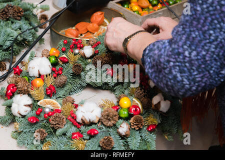 Ghirlanda di Natale atelier di tessitura. Donna mani decorazione ghirlanda di vacanza fatta di rami di abete rosso, coni e varie decorazioni organico sul tavolo Foto Stock