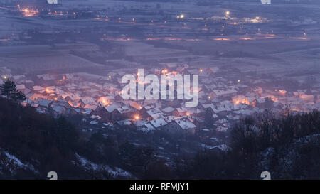 View from above of a dreamy, misty little village covered in snow during late blue hour Foto Stock