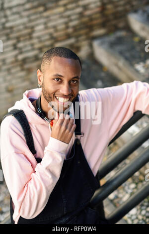Giovane uomo nero indossando abiti casual sorridente su scala urbana. Felice ragazzo africano con bib pants all'aperto Foto Stock