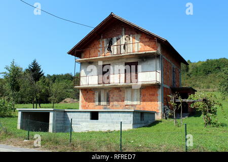 Tall incompiuta abbandonati in mattoni rossi famiglia casa suburbana con imbarcati per porte e finestre, garage incompiuto nella parte anteriore e di erba, alberi e cielo chiaro Foto Stock