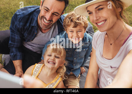 Bella donna con la famiglia sul picnic selfie parlando al parco. Coppia con i loro due bambini facendo un ritratto di auto presso il park. Foto Stock