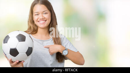 Giovane bella bruna donna azienda soccer football palla su sfondo isolato con sorpresa faccia puntare il dito a se stesso Foto Stock