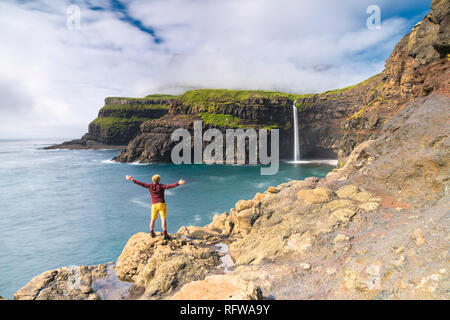 Uomo sulla scogliera con le braccia aperte ammirando Gasadalur cascata, funzionario ministeriale isola, isole Faerøer, Danimarca, Europa Foto Stock