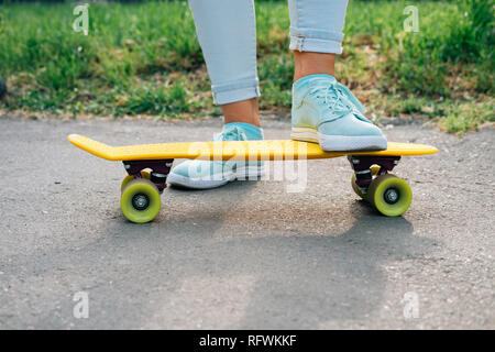 Close-up di donna indossa sneakers in piedi su skateboard sulla soleggiata giornata estiva nel parco vicino a prato verde. Foto Stock