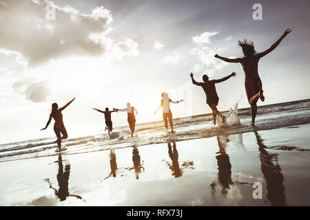 Grande gruppo di amici felice corre e salta al tramonto sulla spiaggia. Sei sagome attivo contro il mare spiaggia Foto Stock