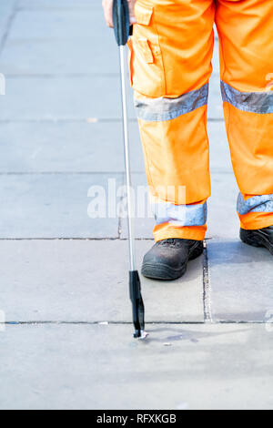 L'uomo lavoratore il detergente in uniforme arancione closeup gambe pulizia nel cestino della spazzatura strade di Londra con strumento di selezione sulla pavimentazione stradale durante il giorno Foto Stock