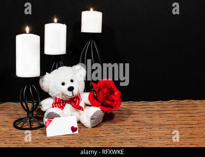 Cudlely orsacchiotto con red bow tie, red rose, candele bianche arroccato su nero portacandele sul posto di maglia mat e tavolo in legno con la carta e scuro bac Foto Stock