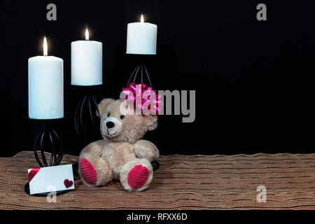 Cudlely orsacchiotto di peluche con fiocco rosa sulla testa, candele bianche arroccato su nero portacandele sul posto di maglia mat e tavolo in legno con la carta e backgrou scuro Foto Stock