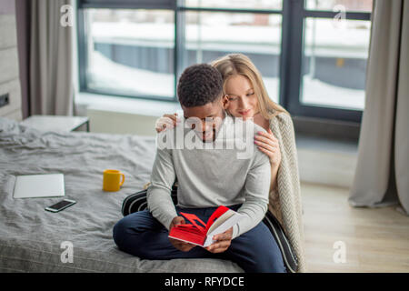 Felice interracial giovane trascorrere weekend insieme a casa, rilassante con libro Foto Stock