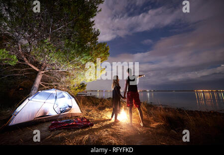 Vista posteriore della coppia giovane e sportiva del uomo e donna slim escursionisti in piedi tenendo le mani vicino a tourist tenda mediante il fuoco. Un uomo indica il cielo di sera e bl Foto Stock