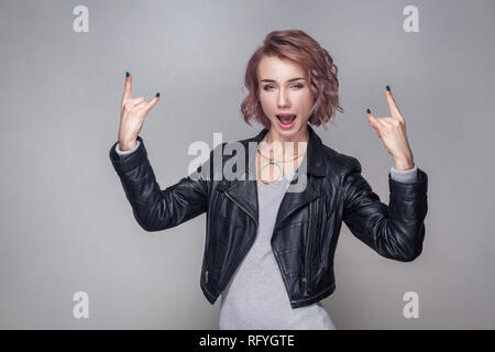 Bella funny stupito ragazza a bilanciere con breve acconciatura e trucco in stile casual nero giacca in pelle in piedi con segno di roccia e guardando la fotocamera. Foto Stock