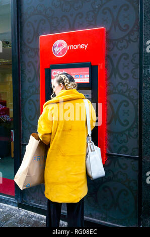 Giovane donna in un rivestimento di colore giallo con una borsa a mano e shopping a una vergine denaro ATM Bancomat Foto Stock