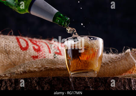 Birra Leggera o Birra Light versa da una bottiglia in un calice di vetro contro lo sfondo di tessili sotto la tela. Close-up. Foto Stock