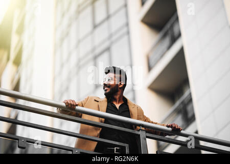 Basso angolo di visione verticale di un uomo indiano indossando occhiali da sole e giacca mentre fantasticando all aperto in una città moderna Foto Stock