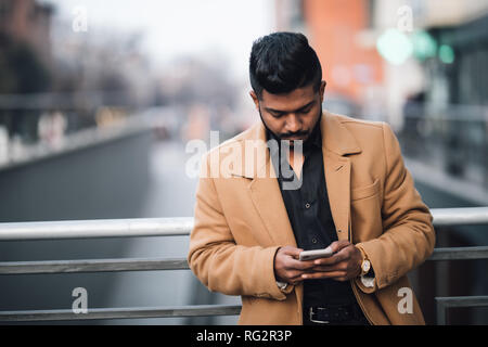 Ritratto di barbuto uomo indiano nel rivestire la digitazione di messaggio sul telefono sulla strada di città Foto Stock