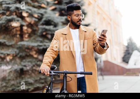 Bello barbuto imprenditore in abito classico è utilizzando uno smart phone e sorridenti mentre cavalcate in bicicletta in città Foto Stock