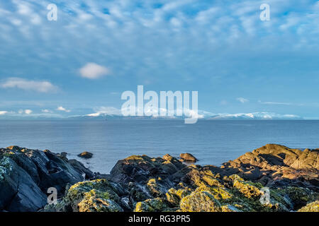 L'isola di Arran in inverni prima neve del 2019 dall'foreshore di Portencross in Scozia con il lichen ricoperta di rocce e la luce riflessa dal w Foto Stock