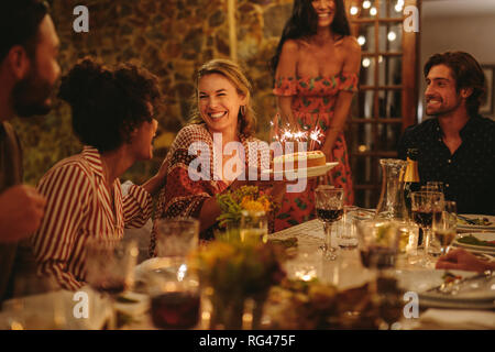 Bella giovane donna festeggia il suo compleanno con i suoi amici. Azienda femmina torta di compleanno seduto con gli amici intorno al tavolo da pranzo all'aperto. Foto Stock