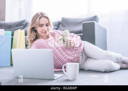 Allegra donna guardando laptop mentre si trattiene le banconote di dollari a casa Foto Stock