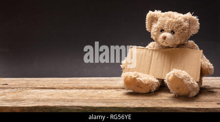 Poveri bambini senzatetto elemosina. Orsacchiotto triste, tenendo un vuoto segno di cartone, seduta in sfondo nero, banner, spazio di copia Foto Stock