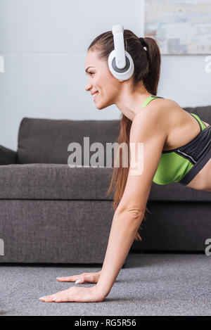 Bella sorridente sportive nelle cuffie facendo plank esercizio a casa Foto Stock