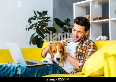 Felice giovane uomo con notebook petting cute cane sul divano Foto Stock