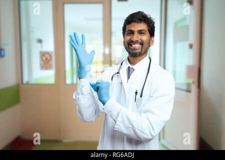 Maschio indiano medico o medic ottenere pronto per la chirurgia come indossare chirurgico blu guanti in lattice all'interno di ospedale Foto Stock