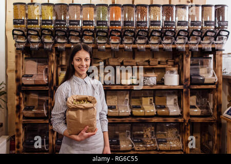 Ritratto del negoziante sorridente nel pacchetto gratuito drogheria. Allegro shop assistant holding sacchetto di carta pieno di erba essiccata foglie in rifiuti zero shop. Foto Stock