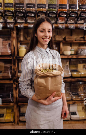 Ritratto del negoziante sorridente nel pacchetto gratuito drogheria. Allegro shop assistant holding sacchetto di carta pieno di erba essiccata foglie in rifiuti zero shop. Foto Stock