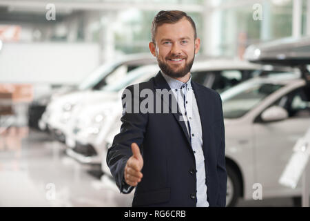 Bello manager della concessionaria auto guardando la telecamera, sorridente, posa. Concessionaria auto indossando in maglietta ufficiale, blu scuro tuta. Felice l'uomo barbuto che mostra il pollice verso l'alto. Foto Stock