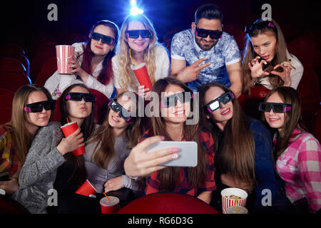 Positività e divertente gruppo di studenti fare foto sullo smartphone e tenendo selfie. Molte belle ragazze in telecamera durante il film in sala cinema tenendo autoritratto di indossare gli occhiali 3d. Concetto di divertimento. Foto Stock