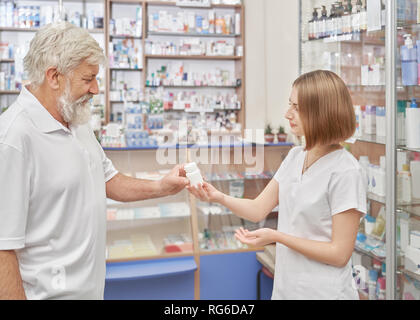 Vecchio cliente di farmacia in consultazione con il farmacista sui  medicinali. Specialista di sesso femminile aiutando, dando pillola bianco  bottiglia e istruire. Titolare di pensione o di rendita con i capelli grigi