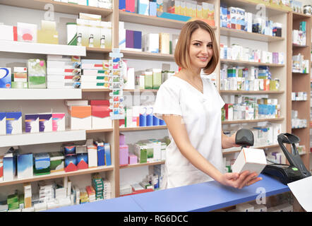 Bella donna che indossa in bianco camice lavora in farmacia. Felice il farmacista permanente al contatore in farmacia. Specialista sorridente, guardando la fotocamera, la scansione di codici a barre medicamento con scanner. Foto Stock