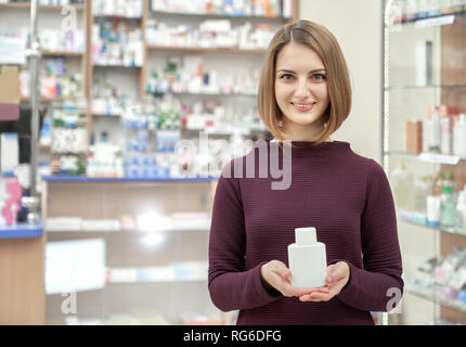 Bella donna di contenimento del prodotto cosmetico in mani. Farmacia cliente sorridendo, guardando la telecamera, posa con bottiglia bianca. Client femmina l acquisto di prodotti in farmacia. Foto Stock