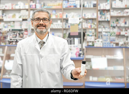 Bello il farmacista che pongono in farmacia, tenendo bottiglia di plastica bianca in mano. Uomo Barbuto guardando la telecamera, sorridenti e in posa. Il farmacista indossa in bicchieri, bianco camice. Foto Stock