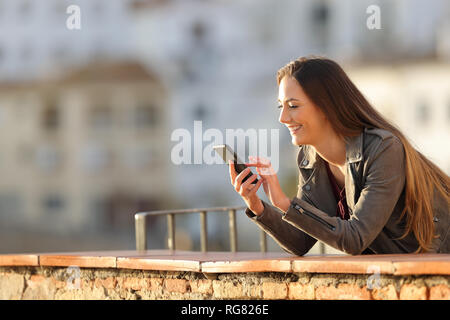 Donna felice utilizza un telefono intelligente in un balcone al tramonto in una città Foto Stock