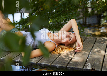 Felice donna bionda giacente sul molo di legno in un lago Foto Stock