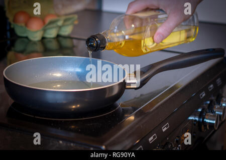 Versare olio di cottura in un tegame in preparazione per preparare la colazione al di sopra di un piano di cottura di nero. Foto Stock