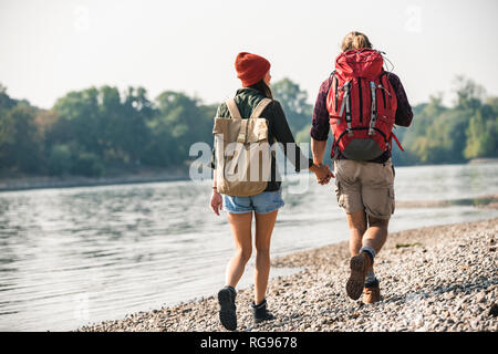 Vista posteriore della giovane coppia con zaini camminando mano nella mano al Riverside Foto Stock