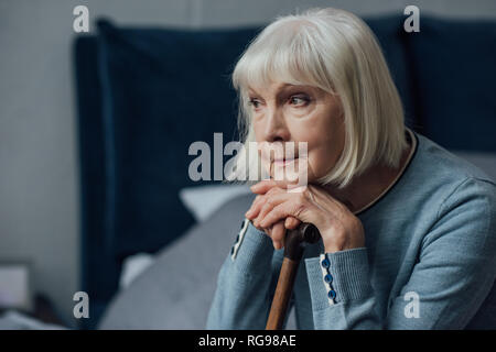 Riflessivo senior donna con le mani sul bastone da passeggio seduta sul letto di casa Foto Stock